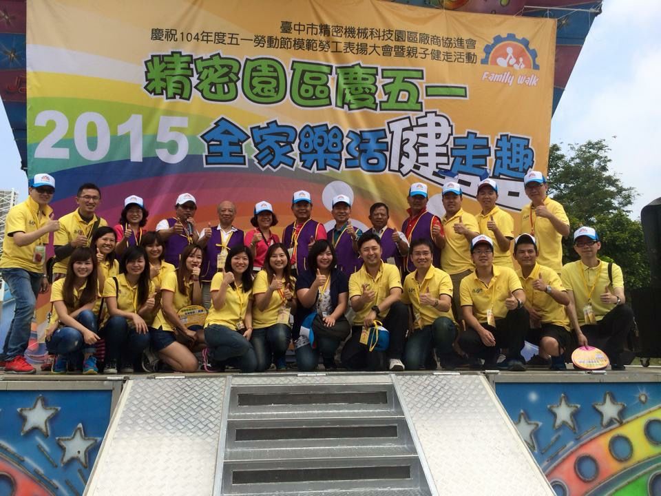 2015 年精密園區全家樂活健走活動，全體人員穿著黃色隊服在舞台前大合照。